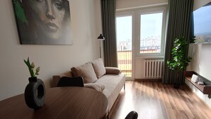 A modern living room with a sofa, a dining table with chairs, a potted plant, and a large window with curtains.