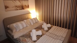 A neatly made bed with pillows and towels, a painting on the wall, and a window with vertical blinds.
