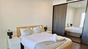 Un lit bien fait, avec une couette blanche et des oreillers, une lampe de chevet, une table de chevet en bois et un grand miroir qui reflète la pièce.