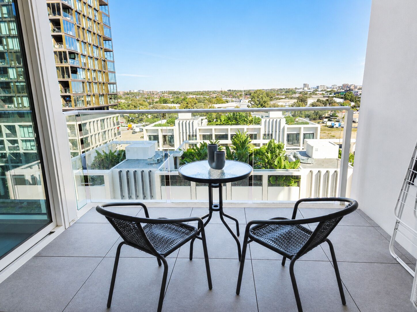 A balcony with two chairs and a small table, overlooking a cityscape with modern buildings and greenery.