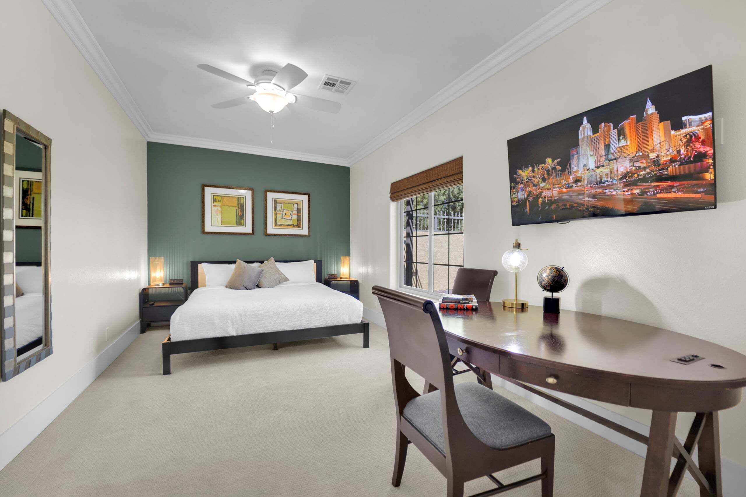 A hotel room with a bed, a desk, a chair, a ceiling fan, framed artwork, and a cityscape painting.