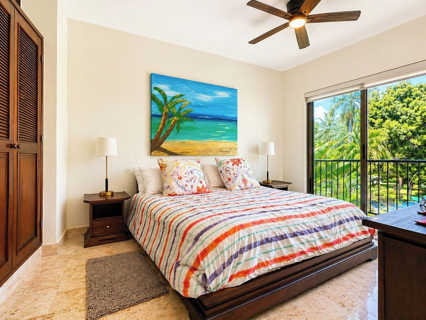 Ein Schlafzimmer mit einem Bett, zwei Nachttischen, einem Deckenventilator, einem Strandbild und einem Fenster mit Blick auf Grün.