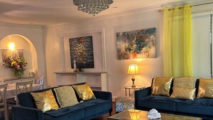 En stue med to blå sofaer, et sofabord i glas, en lysekrone og kunst på væggene.