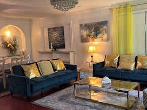 Ein Wohnzimmer mit zwei blauen Sofas, einem Glastisch, einer Pendelleuchte und Kunstwerken an den Wänden.