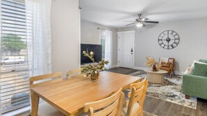 A dining area with a wooden table and chairs, a ceiling fan, a clock on the wall, and a green sofa in the living room.