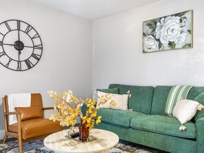 Un sal ón con un sofá verde, un sillón de cuero marrón, una mesa de centro redonda, un reloj de pared y un cuadro con flores.