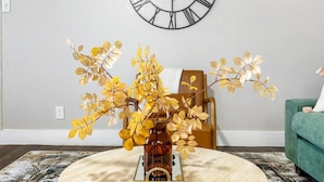 A room with a large wall clock, a vase with yellow leaves, a brown chair, and a green sofa.