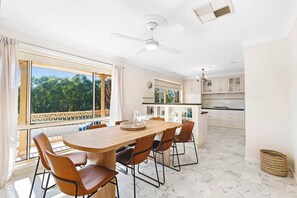 Zona pranzo con un tavolo grande e sedie, una ventola a soffitto e vista sugli alberi attraverso ampie finestre.