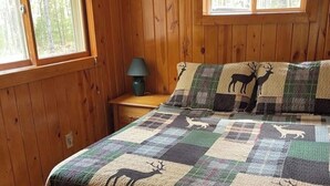 A wooden cabin room with a bed, two windows, a nightstand, and a lamp.