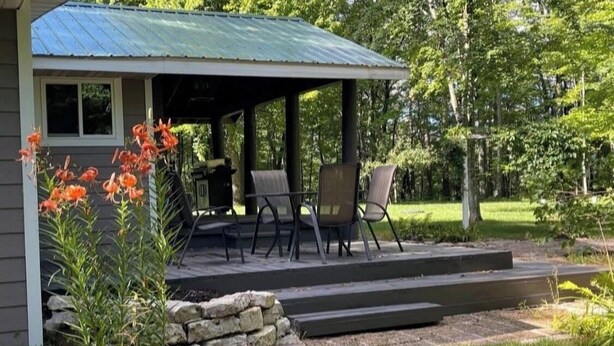 A covered outdoor seating area with chairs and a table, a stone wall, and a building with a metal roof.