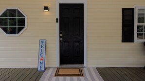 Ett hus med en svart dörr, en välkomstskylt, ett fönster med fönsterluckor och en veranda med en dörrmatta.