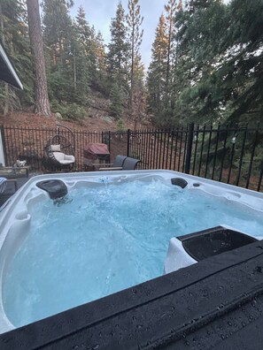 Een hottub omringd door een houten terras, een metalen hek en een bebost gebied.