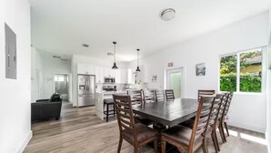 A modern kitchen with white cabinets, a dining area with a large table and chairs, and a view of greenery outside.