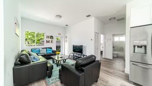 A modern living room with a sofa set, a coffee table, and a flat-screen TV. There is a kitchen area with a refrigerator and a water dispenser.