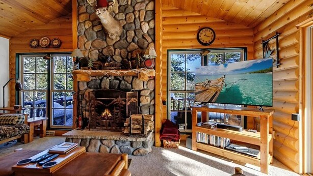 Interior de una cabaña de troncos con chimenea de piedra, muebles de madera, televisor y un ventanal con vistas al exterior.