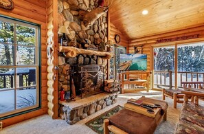 A log cabin interior with a stone fireplace, a television, and a view of a snowy landscape.