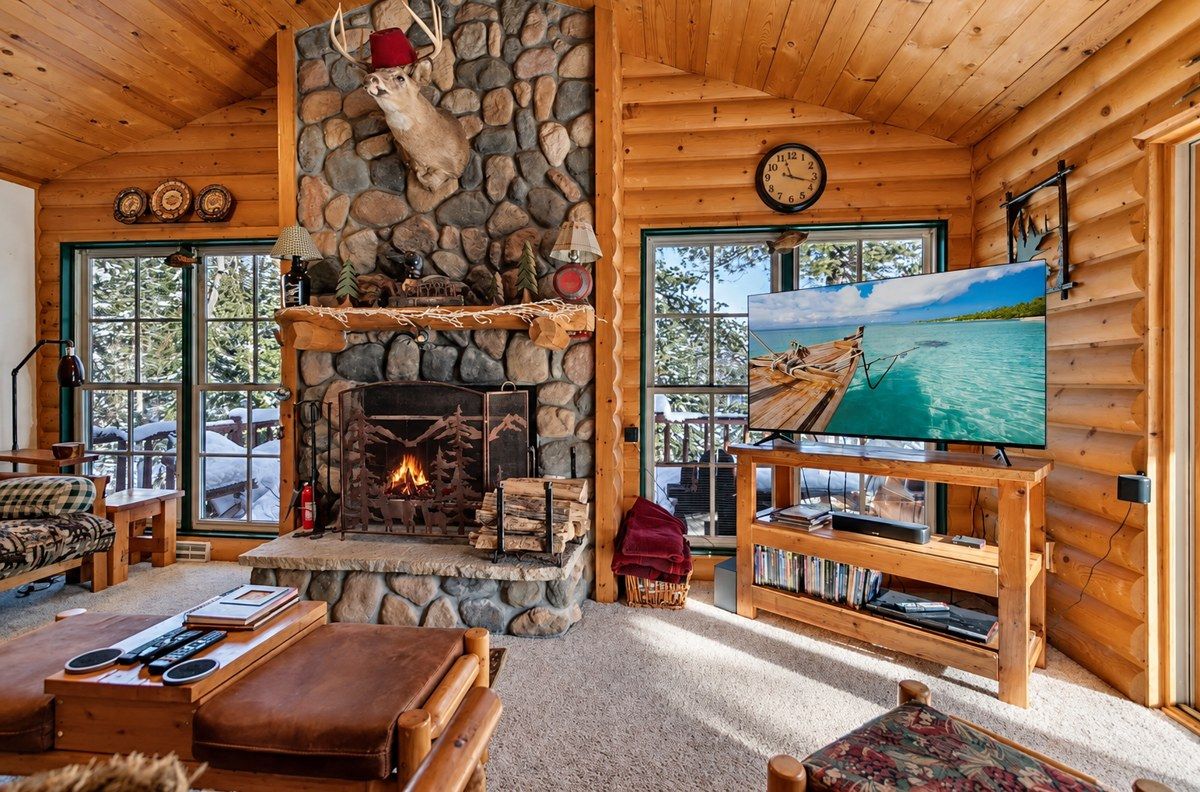 A cozy log cabin interior with a stone fireplace, wooden furniture, and a television displaying a scenic view.