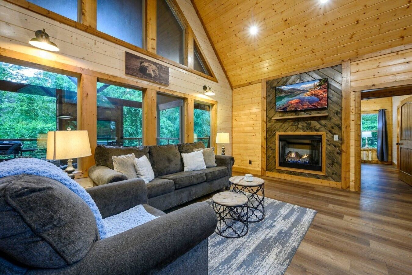 A cozy cabin interior with a fireplace, wooden walls, and large windows overlooking greenery.