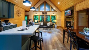 Interno di una baita in legno con cucina, sala da pranzo e soggiorno, caratterizzato da pareti in legno, un caminetto e ampie finestre.