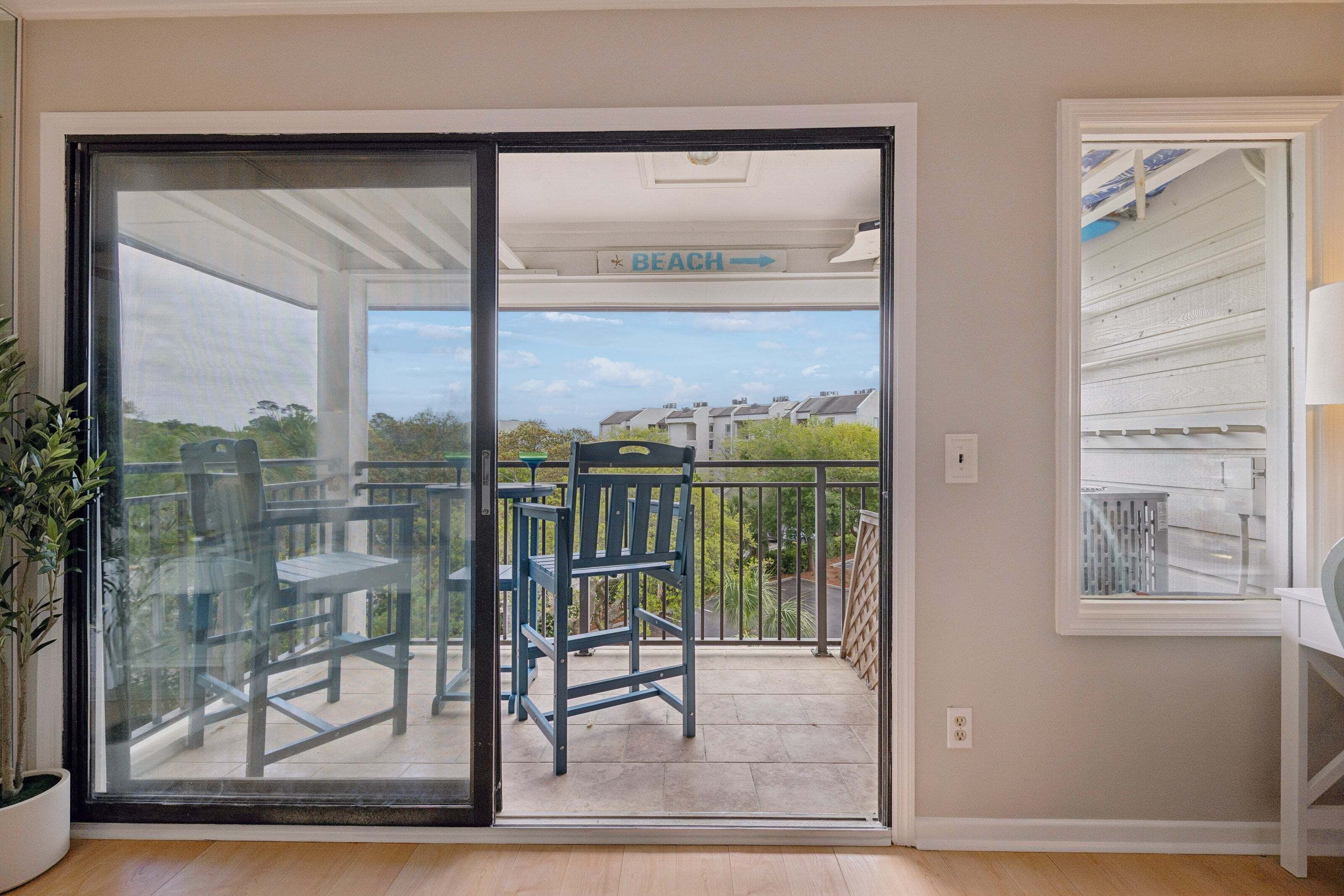 A balcony with a sliding glass door, a chair, a table, and a view of the beach and houses in the distance.