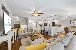 Ein modernes Wohnzimmer mit Sofa, Couchtisch und Fernseher. Es gibt eine Küchenzeile mit weißen Schränken und einem Esstisch.