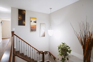 Una escalera con barandilla de madera y balaustres metálicos, una lámpara colgante y obras de arte en la pared.
