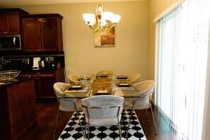 Un comedor con una mesa de vidrio, cuatro sillas, una lámpara de araña y una cocina con armarios de madera oscura y un microondas.