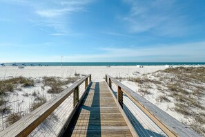 Ein Holzsteg führt zu einem Strand mit Dünen und einem wolkenlosen, blauen Himmel.