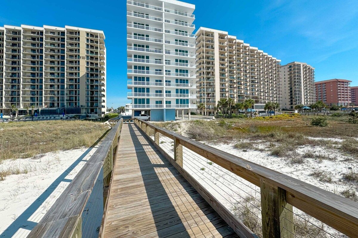 Eine Promenarde führt zu Strandapartements mit mehreren Balkonen, einem Sandstrand und einem klaren Himmel.