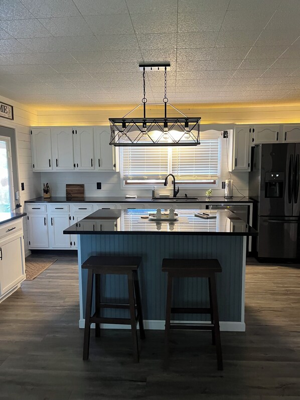 A kitchen with white cabinets, a central island with stools, a hanging light fixture, and a refrigerator.