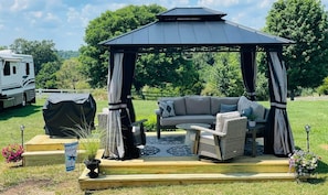 A gazebo with a cushioned seating area, a covered grill, and a welcome sign.