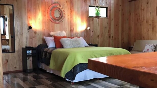 A wooden cabin interior with a bed, a small table, a mirror, and a wall-mounted light fixture.