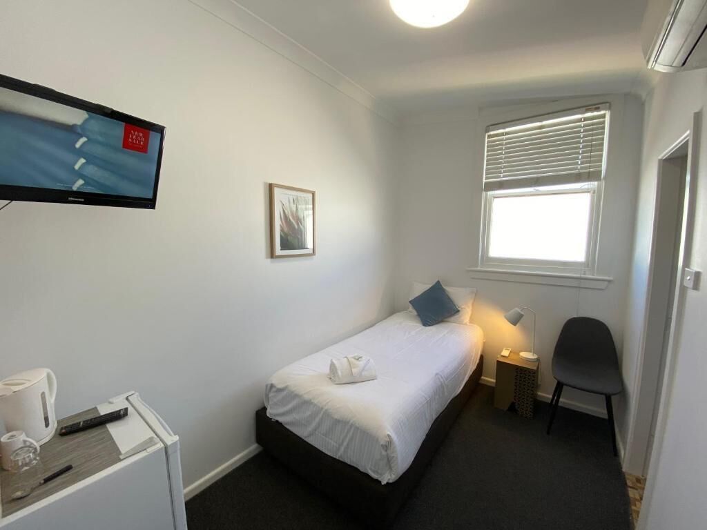 A small, clean room with a single bed, a desk with a chair, a TV, and a window with blinds.