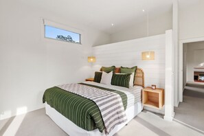 A modern bedroom with a large bed, two bedside tables, a small window, and a view of a staircase. - Bridge Road Retreat – Barwon Heads Getaway (Barwon Heads)