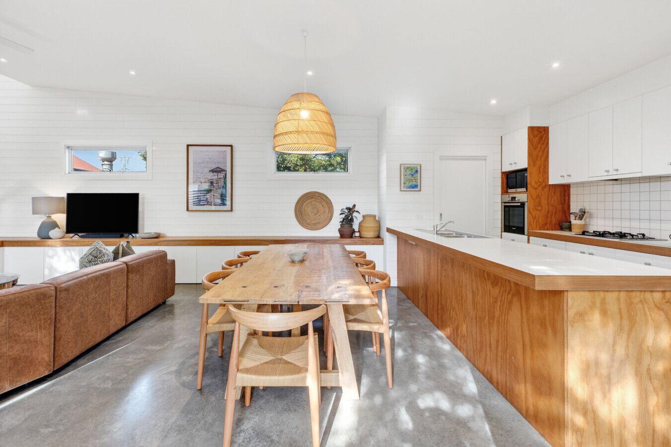 A modern kitchen with a central island, wooden chairs, a dining table, and a TV on a white wall.