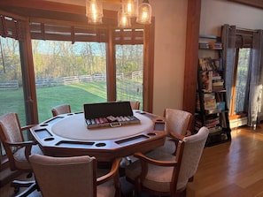 A round wooden table with a laptop, surrounded by cushioned chairs, in a room with large windows and a bookshelf.