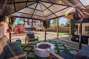 Un patio al aire libre con zona de estar cubierta, un hogar para fuego y una estructura de juegos para niños al fondo.