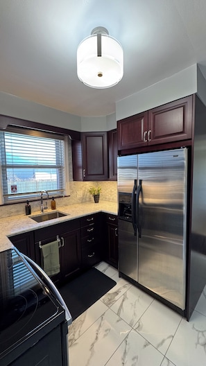 A modern kitchen with dark wood cabinets, a stainless steel refrigerator, a window with blinds, and a marble floor.