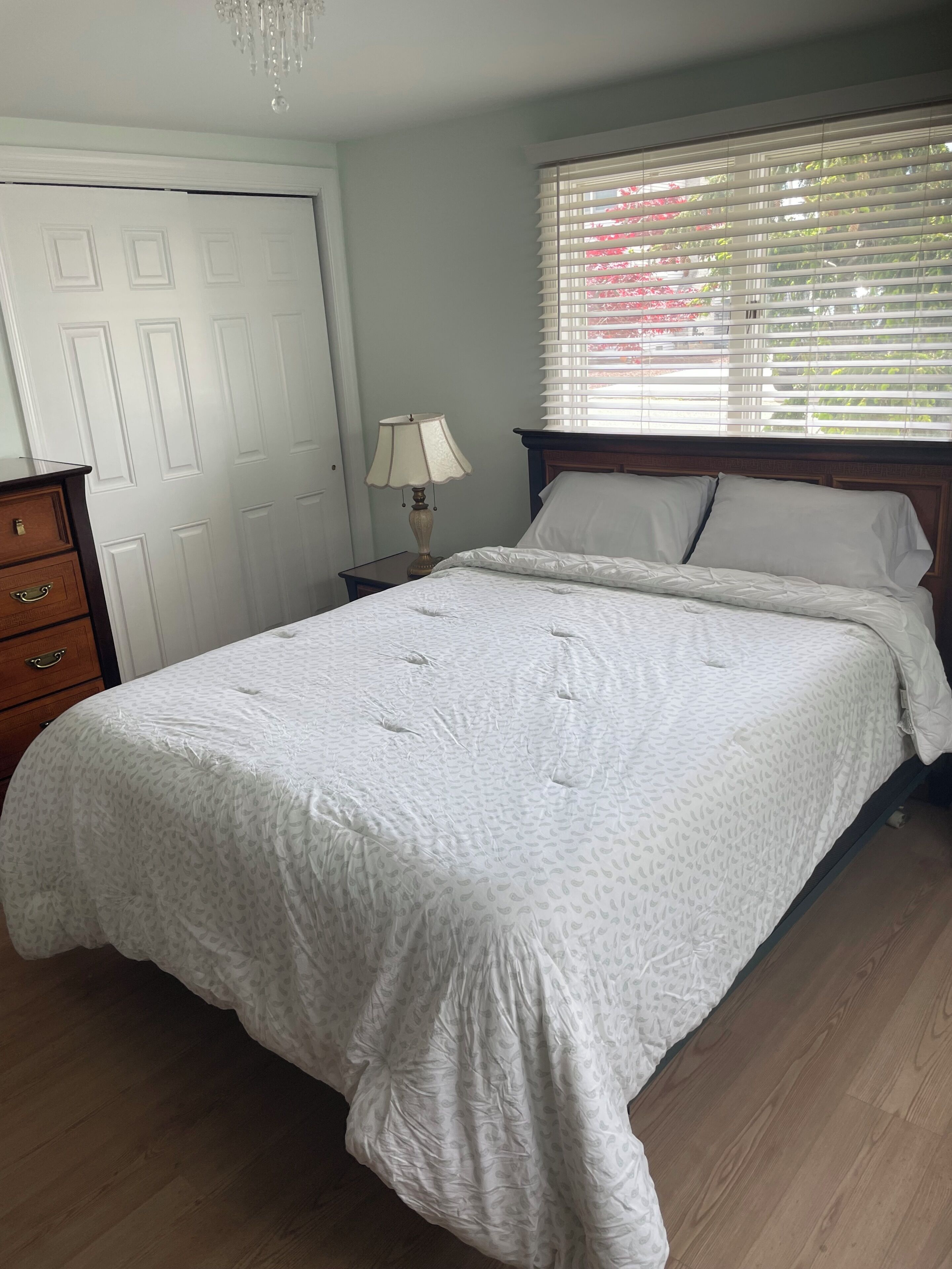 A bedroom with a bed, dresser, nightstand, lamp, and window with blinds.