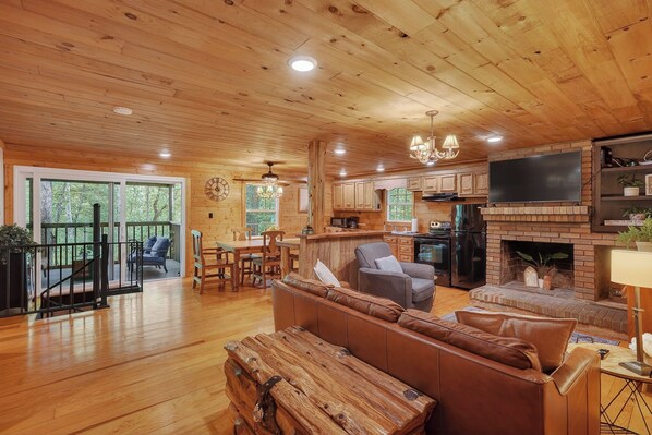 Amplia sala de estar con techo de madera, chimenea, televisor y comedor con vistas al exterior.
