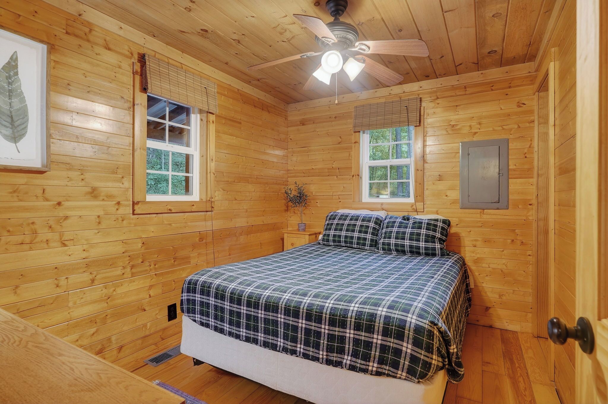 A wooden cabin bedroom with a bed, ceiling fan, two windows, and a door.