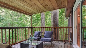 Una terrazza coperta con pavimento in legno, due sedie con cuscini, un tavolo in vetro e vista su alberi fitti.