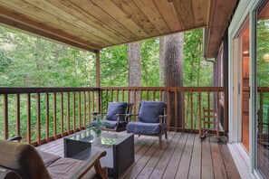 Una terraza techada con suelo de madera, dos sillas con cojines, una mesa de cristal y vistas a árboles frondosos.