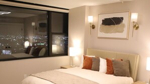 Ein Schlafzimmer mit einem großen Bett, Blick auf die nächtliche Stadt durch ein Fenster und einem an der Wand befestigten Kunstwerk.
