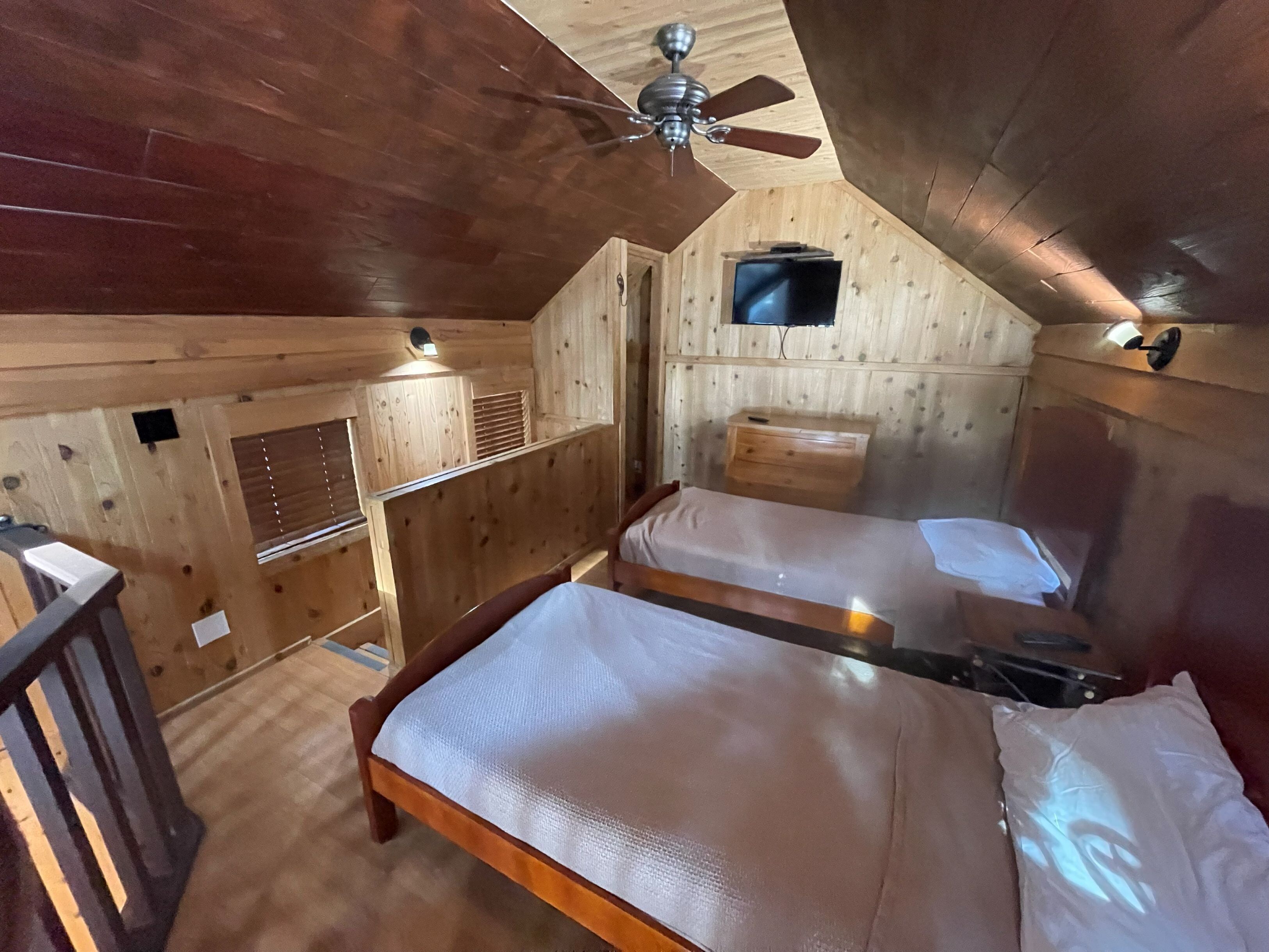 A cabin with two beds, a ceiling fan, a TV, and a wooden interior.