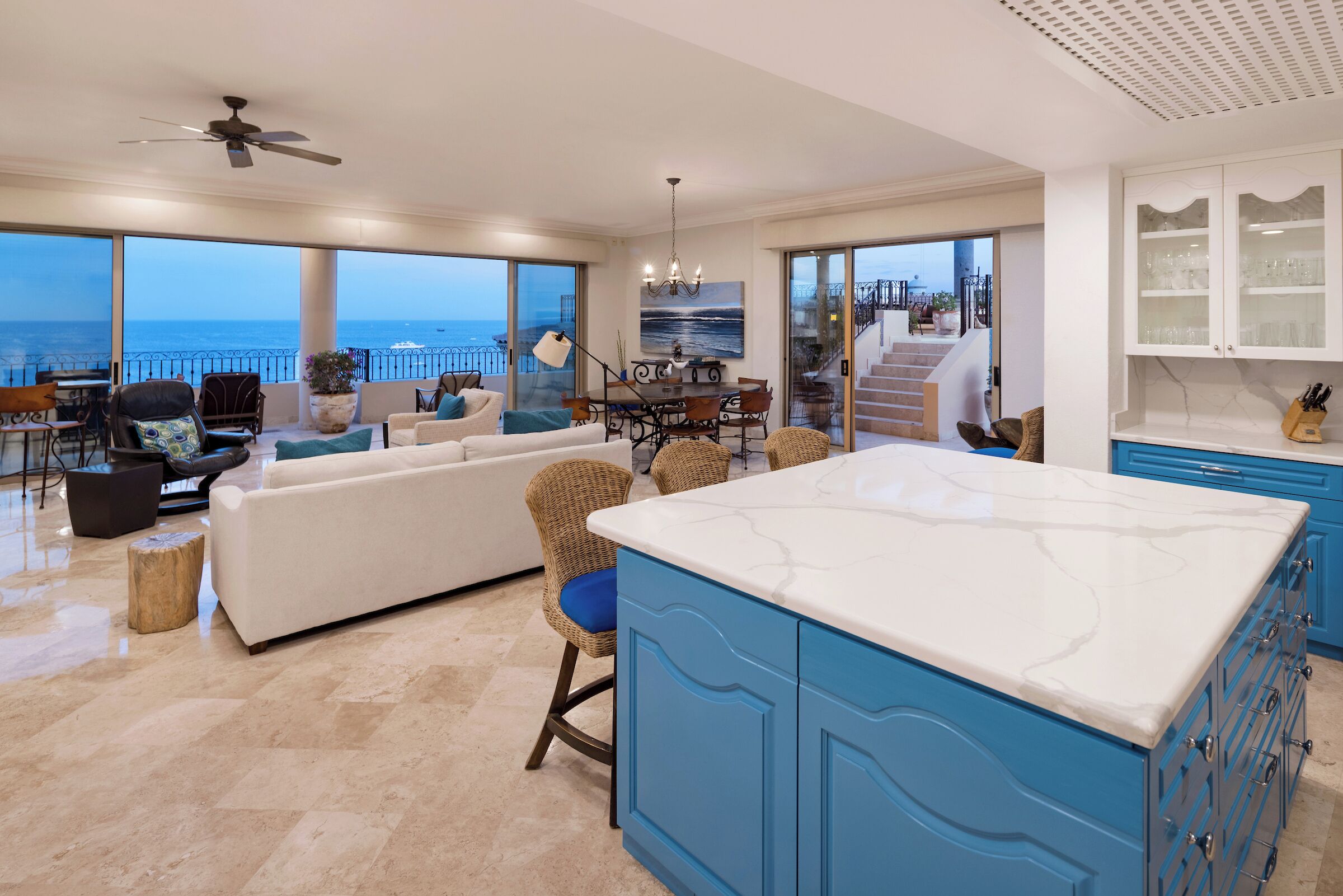 Uma cozinha moderna com armários azuis, bancada de mármore branco e área de jantar com vista para o oceano.