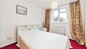 Ett sovrum med en säng, ett sängbord, en lampa, ett fönster med gardiner och ett element.