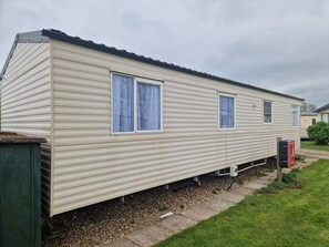 A single-story mobile home with a flat roof, two windows with curtains, and a small attached garage. - Caravan Sleeps 8 Pets Welcome wifi (Skegness)