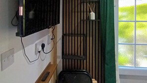 Una habitación compacta con televisor montado, un pequeño espacio de descanso, una ventana con vistas y un estante en la pared con objetos.