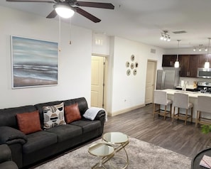 A living room with a sofa, a coffee table, a ceiling fan, and a kitchen area with a refrigerator, microwave, and bar stools. - Modern Comfort in the Heart of Southside (Jacksonville)